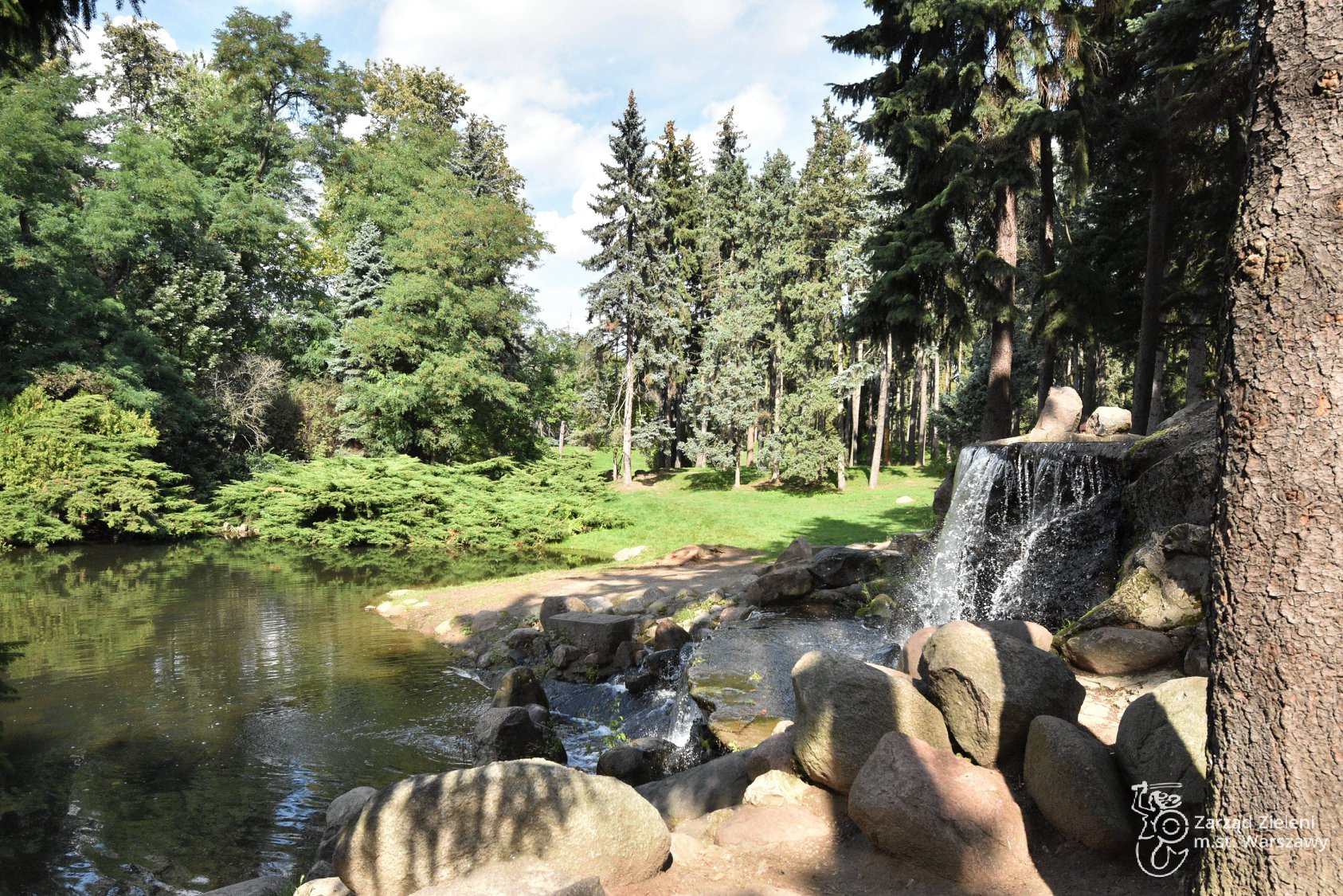Kaskada i staw w Parku Skaryszewskim, woda spływa po skałach, w tle zieleń drzew